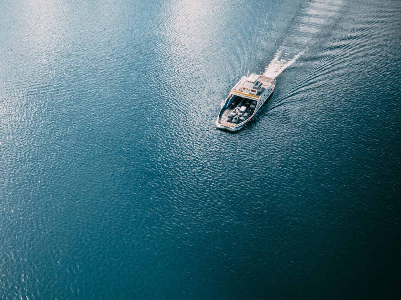 A small passenger ferry crosses open turquoise sea carrying deck cargo and a cool box