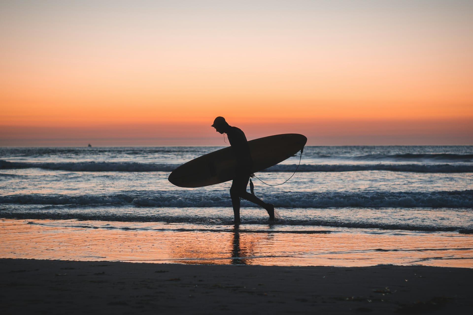 A surfer walks a sandy dirt road through a quiet coastal village at golden hour, board under one arm, palm trees and simple homes behind.