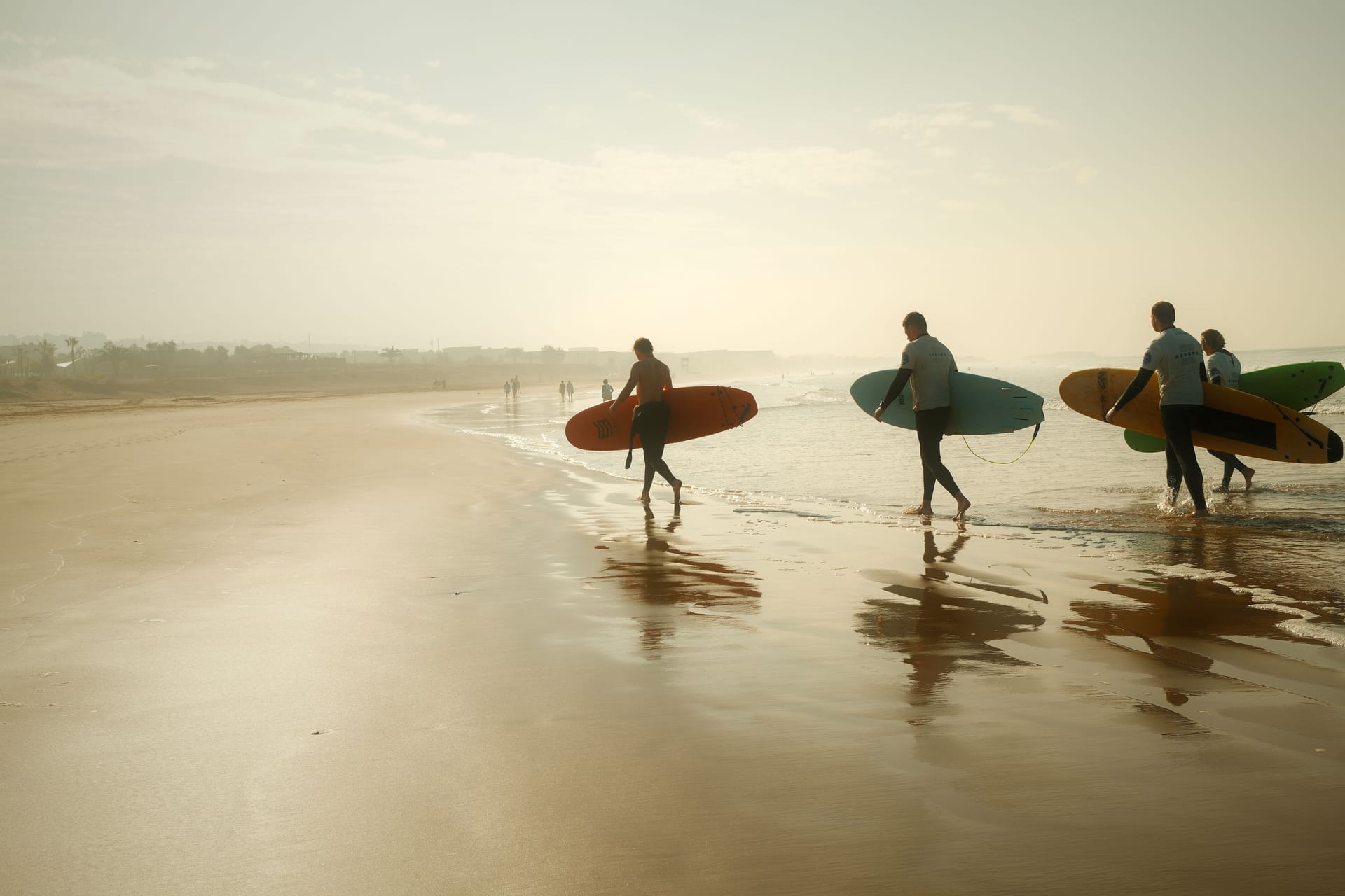 Surfers walking a dirt road through Nemberala village at dawn, with palms and the reef bay behind