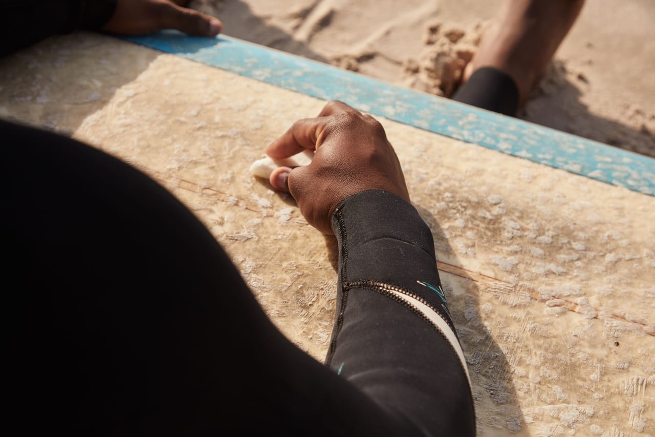 A surfer waxing a board at the water's edge with shallow turquoise reef and exposed limestone behind