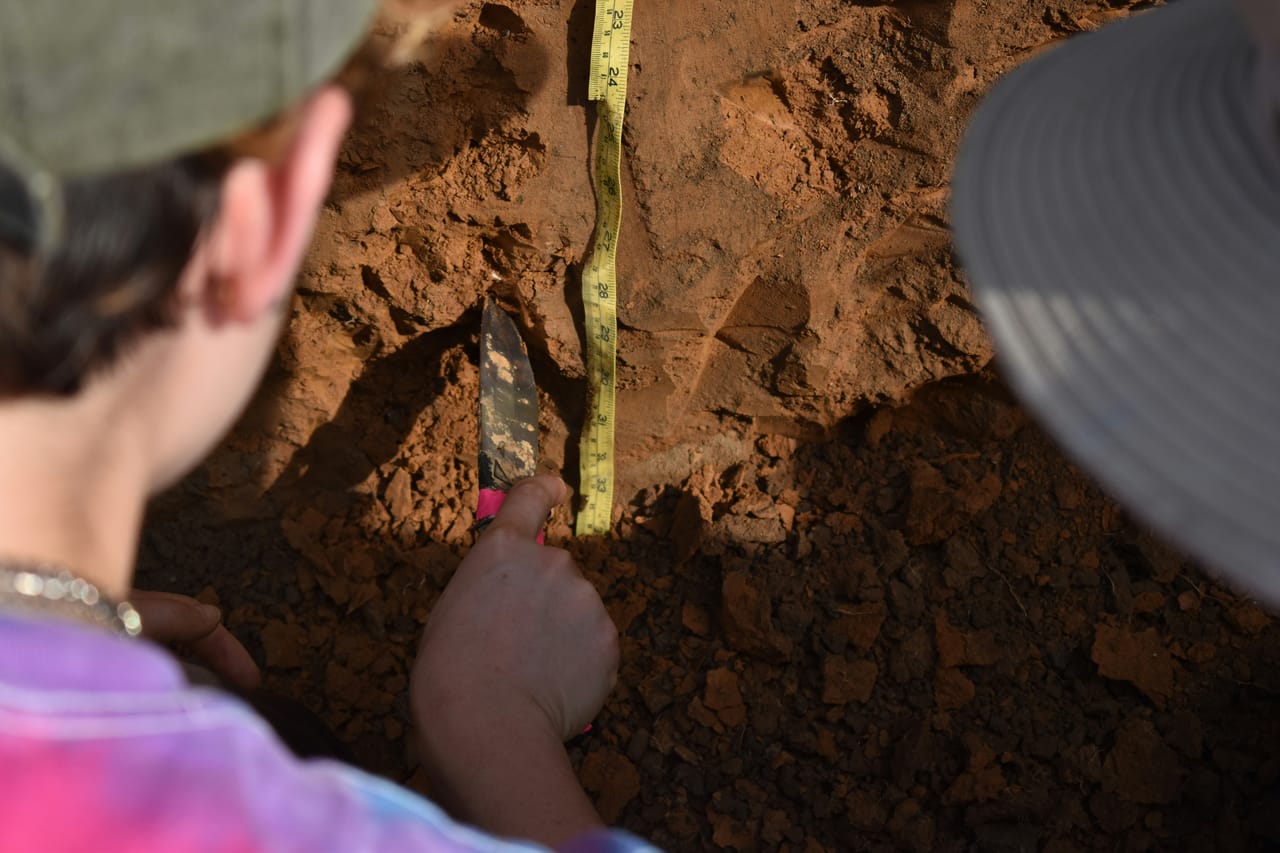 A person crouching on open ground stretching a measuring tape across a plot of bare land