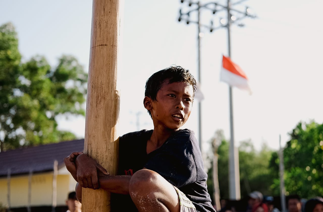 A child climbing a wooden pole in an outdoor setting with the Indonesian flag visible in the background.