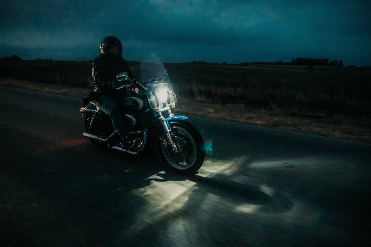 A lone motorbike headlight cutting through a dark, unlit rural road at dusk