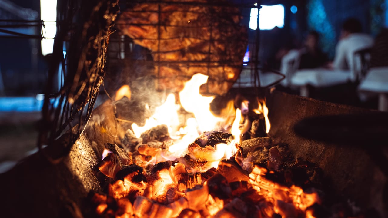 Weathered hands turning strips of meat over embers in a rustic outdoor smokehouse